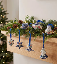 A set of hand-painted ceramic Christmas ornaments styled on a wooden mantel with a green pine garland and warm string lights. The ornaments include a Santa Claus figure, a bird, a star, a heart, a teapot, and a teacup, all decorated with blue, red, and yellow floral patterns on a white base, hanging from blue ribbons.