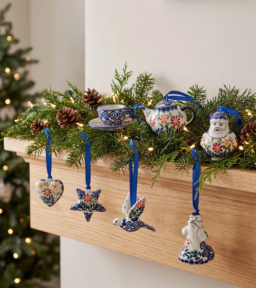 A set of hand-painted ceramic Christmas ornaments styled on a wooden mantel with a green pine garland and warm string lights. The ornaments include a Santa Claus figure, a bird, a star, a heart, a teapot, and a teacup, all decorated with blue, red, and yellow floral patterns on a white base, hanging from blue ribbons.