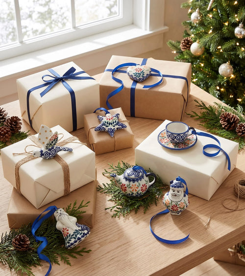 A styled holiday tabletop scene with multiple wrapped gift boxes and decorative porcelain ornaments. The products shown include kraft paper gift wrap, cream gift wrap, blue ribbon bows, floral blue-and-white ceramic ornaments, a small teapot ornament, a Santa ornament, and a teacup-and saucer decoration.