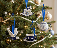 A set of ceramic Christmas tree ornaments in a blue and white polka-dot style, hanging on a decorated Christmas tree. The ornaments include a rocking horse, a teapot, a bird, and a teacup and saucer, each with a blue ribbon loop.