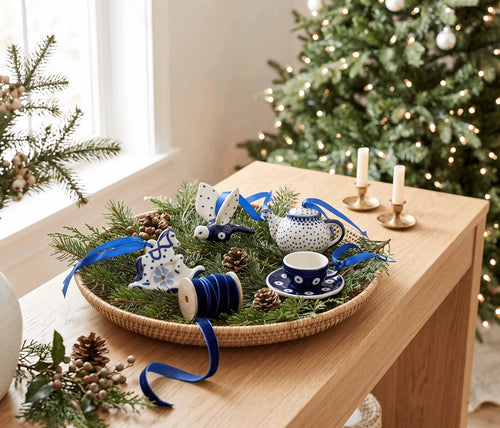 A holiday-themed woven tray filled with green pine branches, blue ribbon, pinecones, and ceramic Christmas decorations including a teapot, teacup, rabbit figurine, and snowman ornament, styled on a wooden table with a decorated Christmas tree in the background.