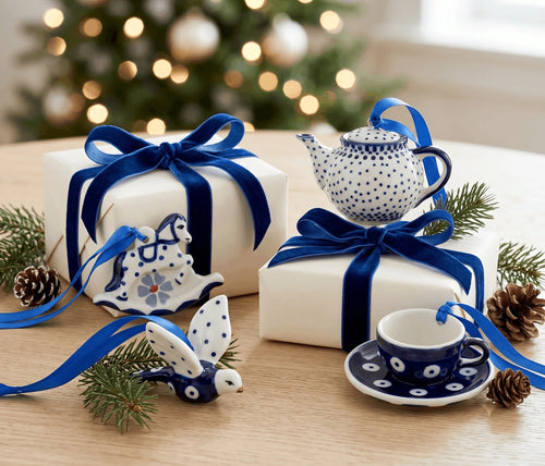 A festive tabletop display of blue-and-white ceramic holiday items, including a teapot, teacup and saucer, ornaments, and wrapped gift boxes with blue ribbons, arranged with pine branches and pinecones in front of a blurred Christmas tree.