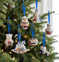 A set of hand-painted ceramic Christmas ornaments shaped like an owl, a rocking horse, a pumpkin, a bear, a Santa figure, and a tractor, hanging on a decorated Christmas tree with blue ribbons.