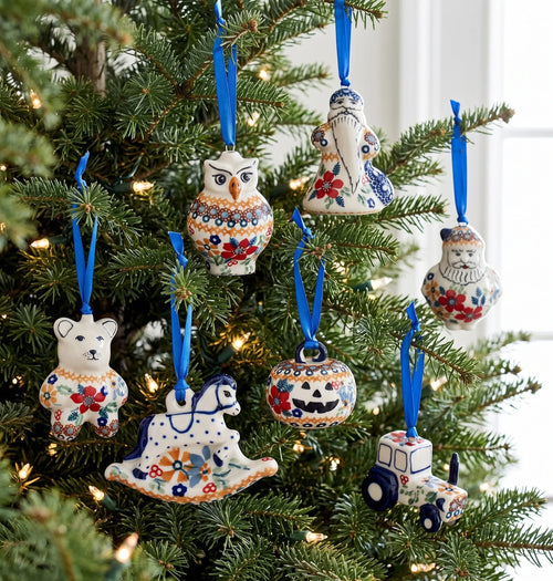A set of hand-painted ceramic Christmas ornaments shaped like an owl, a rocking horse, a pumpkin, a bear, a Santa figure, and a tractor, hanging on a decorated Christmas tree with blue ribbons.