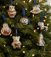 A set of seven hand-painted ceramic Christmas ornaments hanging on a decorated green pine tree with warm string lights. The ornaments feature whimsical winter and farm-themed designs including an owl, a rocking horse, a Santa Claus figure, a jack-o'-lantern, a tractor, a bear, and a cat, all decorated with colorful floral patterns on an off-white background and suspended by blue ribbons.