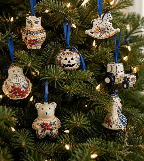A set of seven hand-painted ceramic Christmas ornaments hanging on a decorated green pine tree with warm string lights. The ornaments feature whimsical winter and farm-themed designs including an owl, a rocking horse, a Santa Claus figure, a jack-o'-lantern, a tractor, a bear, and a cat, all decorated with colorful floral patterns on an off-white background and suspended by blue ribbons.