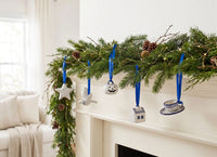 A festive holiday garland draped over a white fireplace mantel in a bright living room. The green pine garland is decorated with small pinecones, white ceramic ornaments shaped like a star, a bird, a snowman, a house, and a teacup, hanging from blue ribbons.