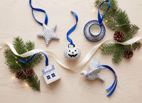 A flat-lay of five hand-painted ceramic Christmas ornaments on a light wood background, decorated with pine branches, pinecones, white ribbons, and fairy lights. The ornaments include a star, a jack-o'-lantern, a house, a bird, and a teacup, in white and blue with black polka dots.