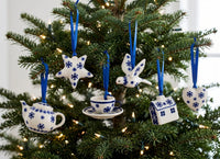 A set of five white and blue ceramic Christmas ornaments shaped like a teapot, teacup, bird, house, and star, hanging on a decorated Christmas tree with warm lights.
