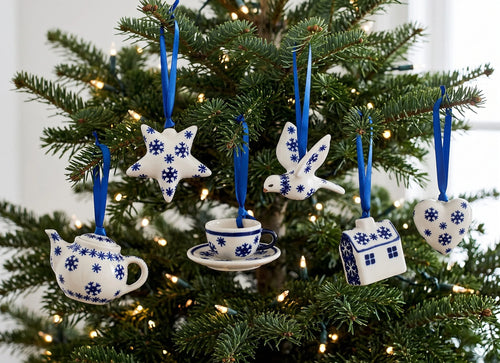 A set of five white and blue ceramic Christmas ornaments shaped like a teapot, teacup, bird, house, and star, hanging on a decorated Christmas tree with warm lights.