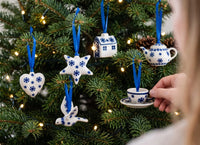 A person's hand hangs a small blue-and-white ceramic Christmas ornament shaped like a teacup and saucer on a decorated evergreen tree with warm string lights. Other hanging ornaments include a heart, star, bird, house, and teapot, all in a matching blue-and-white pattern.