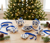 Blue and white ceramic Christmas ornament set on a wooden table with a blurred Christmas tree in the background. The set includes a teapot, cup and saucer, house, bird, star, and heart-shaped ornaments tied with blue ribbons.
