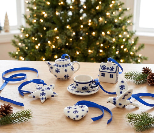 Blue and white ceramic Christmas ornament set on a wooden table with a blurred Christmas tree in the background. The set includes a teapot, cup and saucer, house, bird, star, and heart-shaped ornaments tied with blue ribbons.