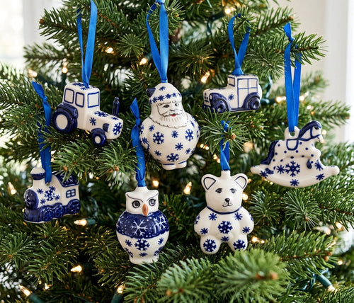 A set of seven blue and white ceramic Christmas tree ornaments shaped like a tractor, Santa, cars, polar bear, owl, and rocking horse, hanging on a green Christmas tree with warm lights.