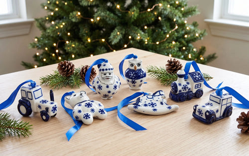 A set of ceramic Christmas ornaments shaped like a Santa Claus, owl, snowman, snowman face, snowman body, snowman hat, snowman boots, snowman hat, snowman boots, and a train, decorated in a blue and white pattern with blue ribbons, arranged on a wooden table in front of a decorated Christmas tree.