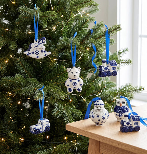 A set of ceramic Christmas tree ornaments in a blue and white Delft style, hanging on a green Christmas tree and displayed on a wooden table. The ornaments include a rocking horse, a teddy bear, a train, a car, a Santa Claus figure, and an owl, each with a blue ribbon hanger.