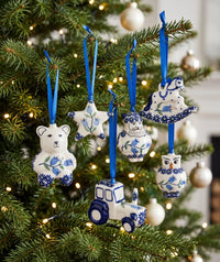 Five ceramic Christmas tree ornaments shaped like a bear, star, flower, rocking horse, and tractor, decorated in a blue and white Delft style with blue ribbons hanging on a decorated Christmas tree.