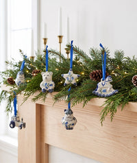Blue and white ceramic Christmas ornaments hanging from a pine garland on a wooden mantel, with white taper candles in the background.