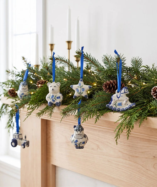 Blue and white ceramic Christmas ornaments hanging from a pine garland on a wooden mantel, with white taper candles in the background.