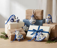 A styled arrangement of wrapped gift boxes and decorative ceramic ornaments in a blue-and-white floral motif, set on a light wood surface with greenery and pinecones against a soft neutral background.