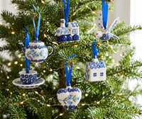 A set of blue and white ceramic Christmas ornaments hanging on a decorated Christmas tree. The ornaments include a teapot, a teacup on a saucer, a bird, a house, a train, and a heart-shaped ornament, all decorated with blue floral patterns on white backgrounds and hanging from blue ribbons.