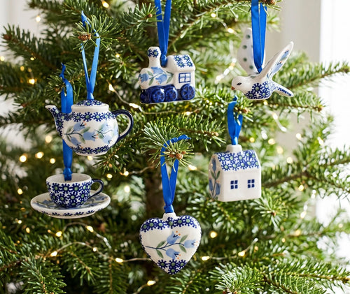 A set of blue and white ceramic Christmas ornaments hanging on a decorated Christmas tree. The ornaments include a teapot, a teacup on a saucer, a bird, a house, a train, and a heart-shaped ornament, all decorated with blue floral patterns on white backgrounds and hanging from blue ribbons.