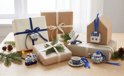 A styled holiday gift arrangement on a wooden table with wrapped presents, blue-and-white ceramic decorative items, pine branches, and a small house-shaped ornament hanging from a wrapped box.