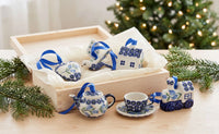 A set of small ceramic Christmas ornaments in a light wood gift box with tissue paper, plus matching teapot, cup, saucer, and a small train ornament displayed on a tabletop with evergreen branches and a blurred Christmas tree in the background.