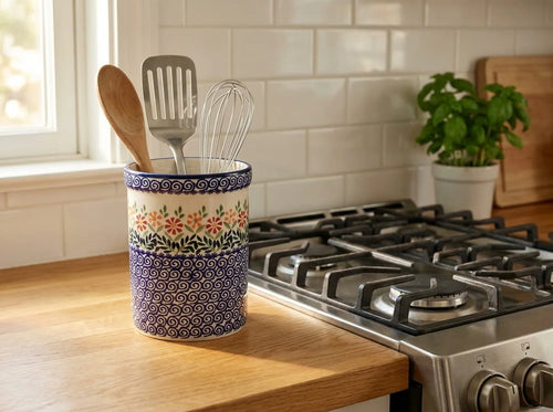 A ceramic kitchen utensil holder with a floral and geometric pattern, containing a wooden spoon, metal spatula, and whisk, sitting on a wooden countertop in a kitchen next to a gas stove.