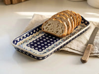 A rectangular ceramic serving tray with a blue and white floral pattern, holding sliced artisan bread and a knife on a beige cloth over a white table.