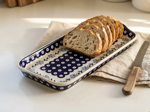 A rectangular ceramic serving tray with a blue and white floral pattern, holding sliced artisan bread and a knife on a beige cloth over a white table.