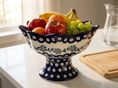 A ceramic pedestal fruit bowl with a white base and navy blue floral patterns, filled with red apples, oranges, green grapes, and bananas, set on a white countertop.