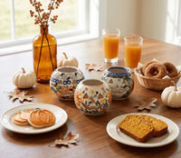 Three decorative ceramic canisters with autumn-themed patterns are arranged on a wooden table alongside fall decorations, plates of food, and glasses of orange juice near a window.