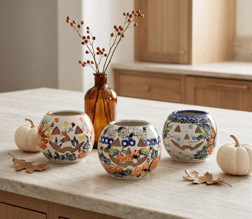 Three decorative ceramic vases with autumn-themed jack-o'-lantern patterns are arranged on a light marble countertop in a kitchen setting, flanked by small pumpkins and a brown glass bottle with dried branches.