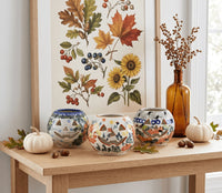 Three hand-painted ceramic pumpkins with carved jack-o'-lantern faces are arranged on a wooden table in a styled home setting, with autumn leaves, acorns, small pumpkins, a tall amber glass bottle, and a framed botanical print in the background.