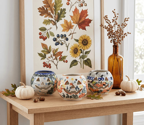 Three hand-painted ceramic pumpkins with carved jack-o'-lantern faces are arranged on a wooden table in a styled home setting, with autumn leaves, acorns, small pumpkins, a tall amber glass bottle, and a framed botanical print in the background.