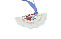 A white ceramic Christmas ornament shaped like a scalloped shell, decorated with red berries, green leaves, and a gold border, hanging from a blue ribbon on a white background.