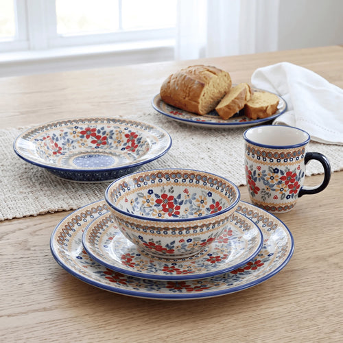 A set of ceramic tableware with a floral pattern in red, blue, and cream, arranged on a light wood table with bread in the background.