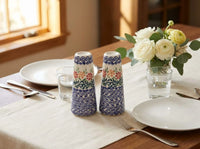 Two decorative ceramic salt and pepper shakers with blue and white scrollwork and floral motifs, set on a dining table next to white plates, cutlery, a glass of water, and a vase of white flowers.