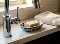 A rectangular bar of soap sits in a small floral-patterned ceramic soap dish on a white bathroom countertop next to a chrome faucet, with a folded beige towel in the background.
