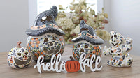 A set of ceramic Halloween decorations on a wooden table, including two witch hat jack-o'-lanterns, a small jack-o'-lantern, a skeleton figure, and a white 'hello fall' sign.