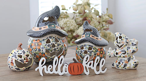A set of ceramic Halloween decorations on a wooden table, including two witch hat jack-o'-lanterns, a small jack-o'-lantern, a skeleton figure, and a white 'hello fall' sign.
