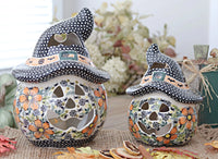 Two hand-painted ceramic witch hat jack-o'-lantern candle holders on a burlap surface with autumn leaves and dried citrus slices, set against a softly blurred background.