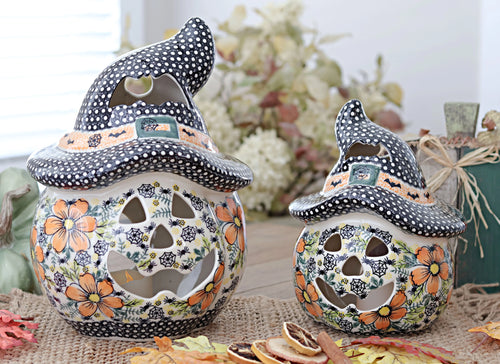 Two hand-painted ceramic witch hat jack-o'-lantern candle holders on a burlap surface with autumn leaves and dried citrus slices, set against a softly blurred background.