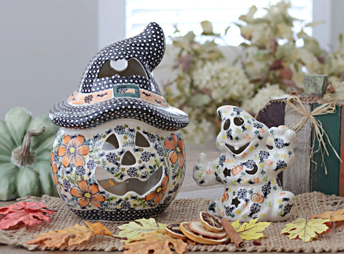 A ceramic Halloween decoration set featuring a jack-o'-lantern-shaped jar with a witch hat lid and a matching ghost figurine, both decorated with floral and spiderweb patterns, displayed on a burlap surface with autumn leaves and a small pumpkin.