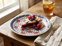 A slice of berry pie on a white ceramic plate with a blue and red floral pattern, set on a rustic wooden table next to a glass of iced tea and a folded napkin with a fork.