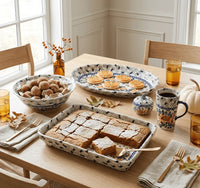A styled dining table set with multiple ceramic dishes and mugs featuring a blue floral pattern on a cream background. The table is set with a large rectangular dish of square brownies, an oval platter of cookies, a bowl of sugar cookies, amber glass cups, a small pumpkin, and autumn leaves on a light wood table in a bright room.