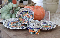 A set of ceramic tableware with a hand-painted floral and pumpkin pattern, displayed on a rustic wooden table alongside decorative pumpkins and a ceramic pot.