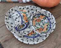A set of white ceramic tableware with a black spider print and orange jack-o'-lantern motifs, arranged on a wooden surface with a decorative pumpkin in the background.