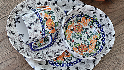 A ceramic tableware set with a white base decorated with black spiderwebs and black spiders, featuring orange jack-o'-lanterns and blue floral borders, shown on a wooden surface.
